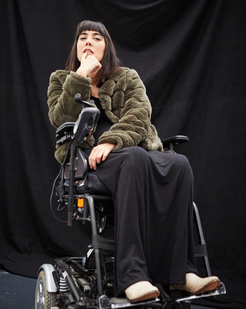 Woman sitting in an electric wheelchair, wearing a green jacket and long black dress.