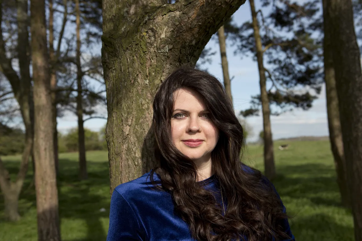 Woman standing near a tree. She has long brown hair and is wearing a blue top.
