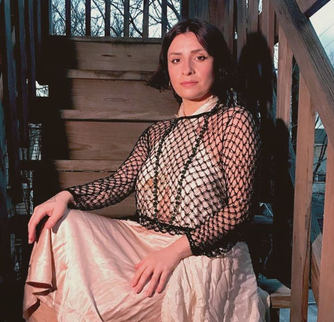 A pale, black haired person with bob sits on wooden stairs in a white dress with a black, fishnet long-sleeve top over it.. 