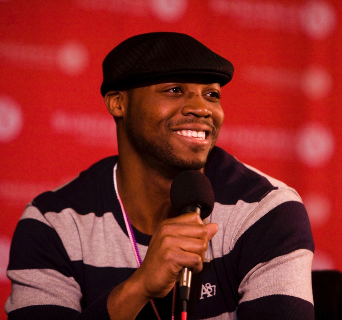 Headshot of an adult African American male wearing a striped sweater and a Kangol hat, holding a microphone in front of red background.