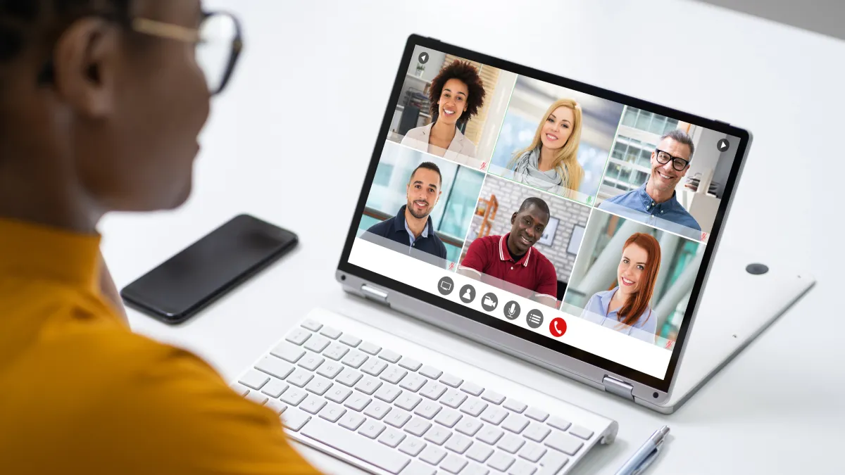 A person wearing a gold shirt is looking at a laptop with six faces on the screen having a virtual meeting. 