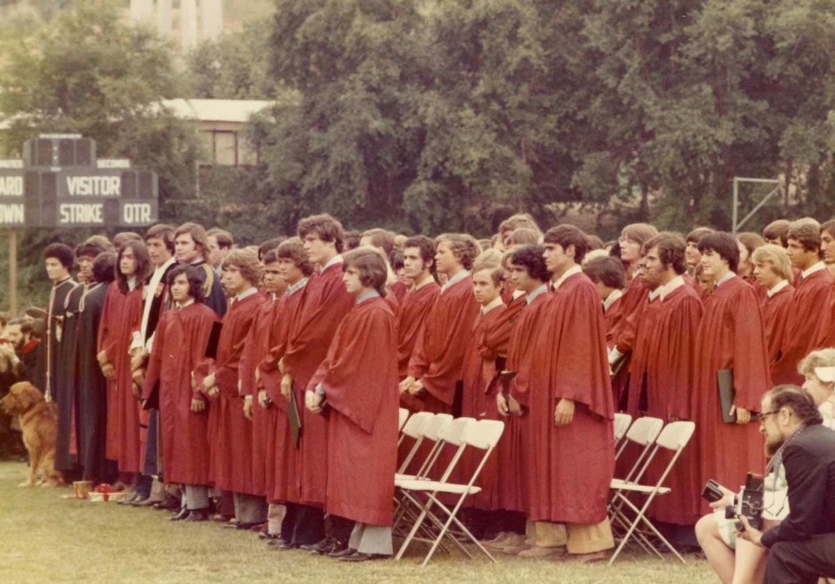 A group of several dozen young men stand on a field wearing red graduation robes. They are holding diplomas.  