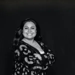 A woman with long, shiny black hair smiles at the camera, her arms crossed. The image is black and white and she wears a patterned cardigan. 