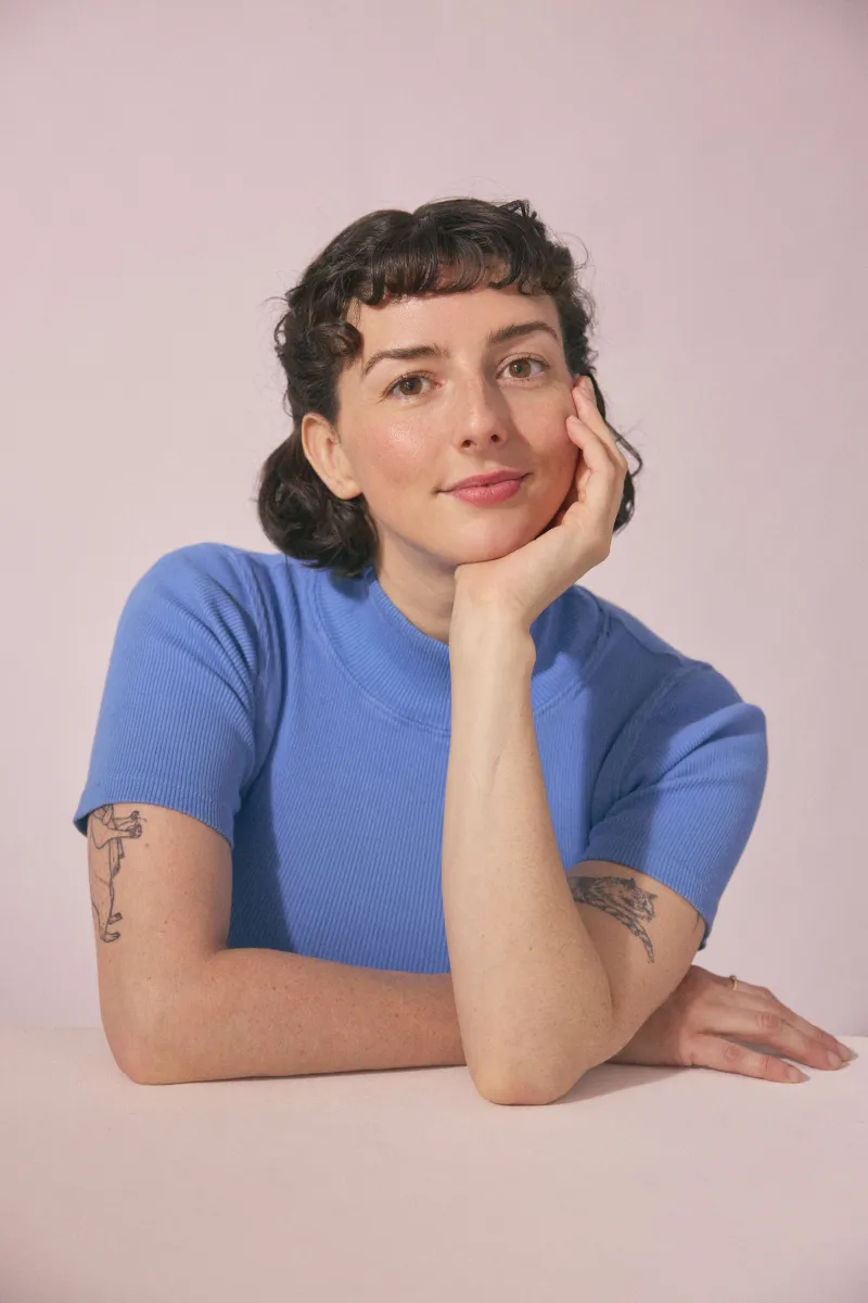A woman with short brown curly hair sits at a table in a blue short sleeve short. She holds one hand on her face and one hand on the counter.
