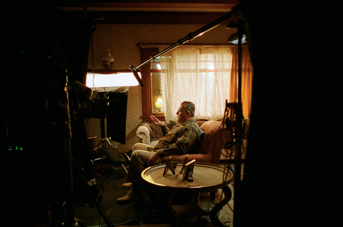 A wide angle shot of a Latino man seated in a living room surrounded by documentary interview equipment.