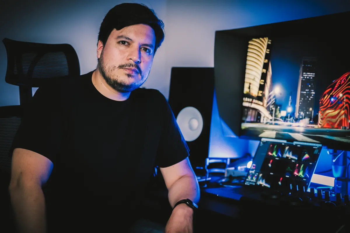 A man sitting in front of a color grading suite looks at the camera. He has short black hair and brown eyes and is wearing a black T-shirt.