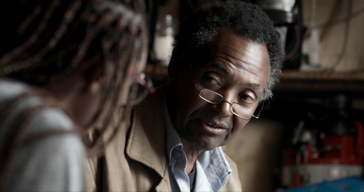An older Black man wearing glasses is interviewing a blurry figure in the foreground