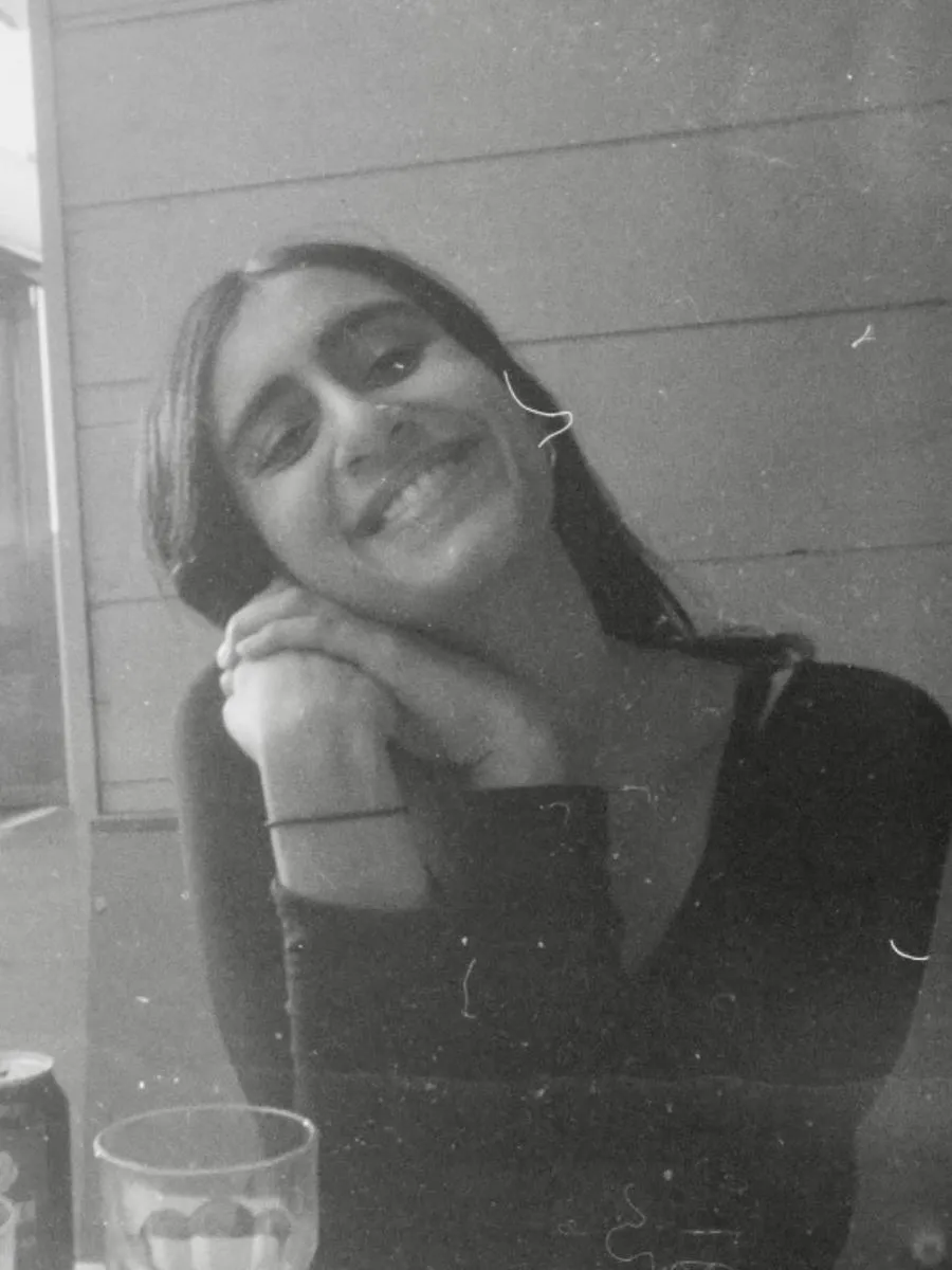 Black and White image of a woman with brown skin smiling at the camera with her head tilted. A woman with white skin smiles at the camera. She has mid-length, straight black hair and is wearing a black top.