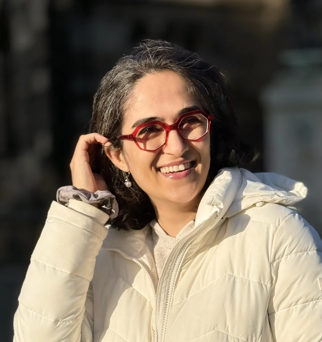 A relatively happy woman in a white jacket, wearing red glasses, brown skin, and short hair, trying to look happier by using her hand to hold her black hair behind her ears.