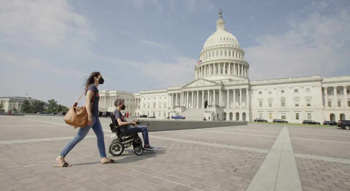 Two people outside a government building wearing masks. One person is in a wheelchair.