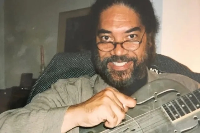 A man with dark hair and a beard, wearing glasses, smiles at the camera while holding a string instrument