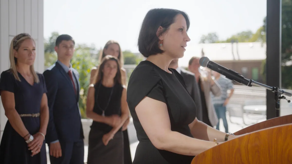 white woman standing at podium in black dress addressing crowd with people standin behind her