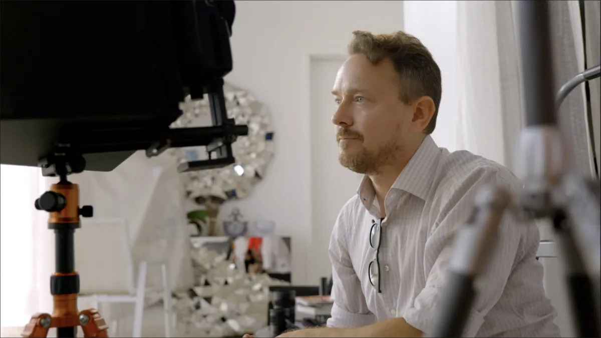A white man sits behind some camera equipment in a dressing room.