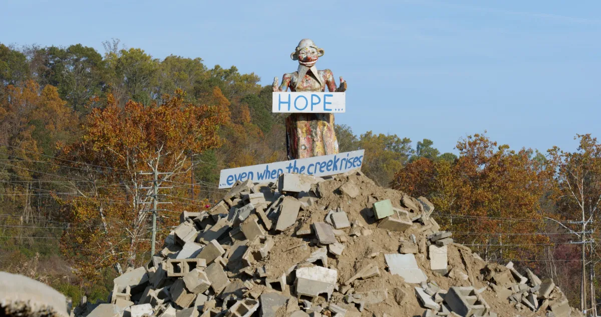 Atop a pile of concrete rubble, there is a statue of a person holding a sign that says "Hope... Even when the creek rises..."