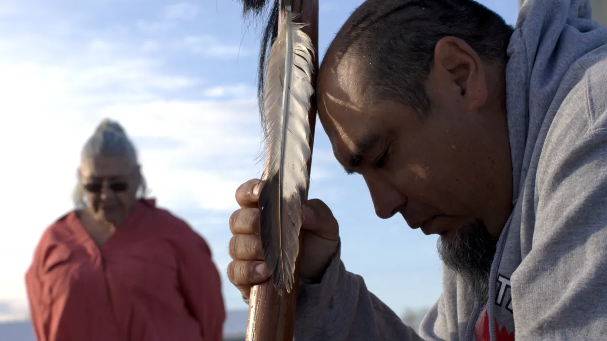 Close up of a man praying, with a woman standing in the background.