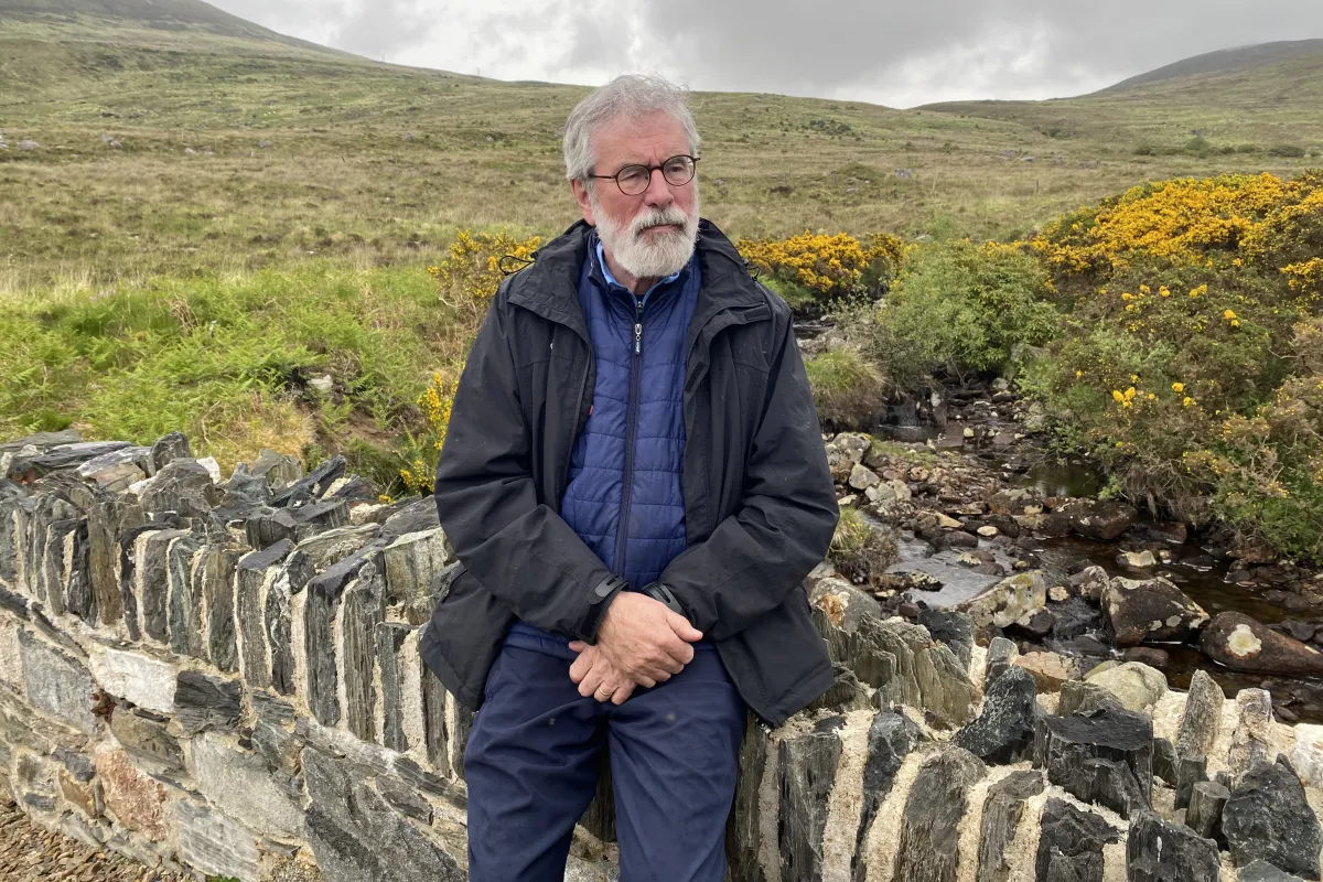 An older man with white hair and a beard, wearing glasses and a jacket, stands in front of a green field and hills. 