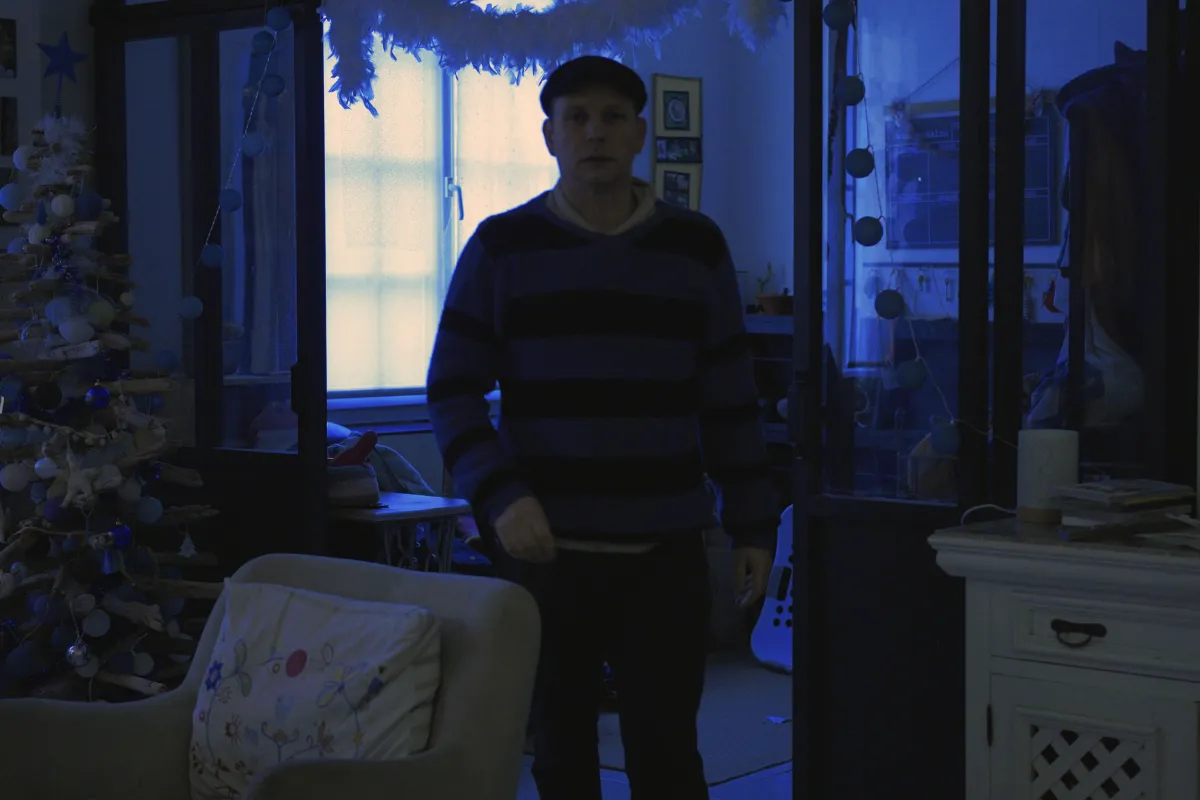 A man in a striped sweater and cap stands in a dimly lit room bathed in a cold blue tint, captured in the early morning light of winter. 