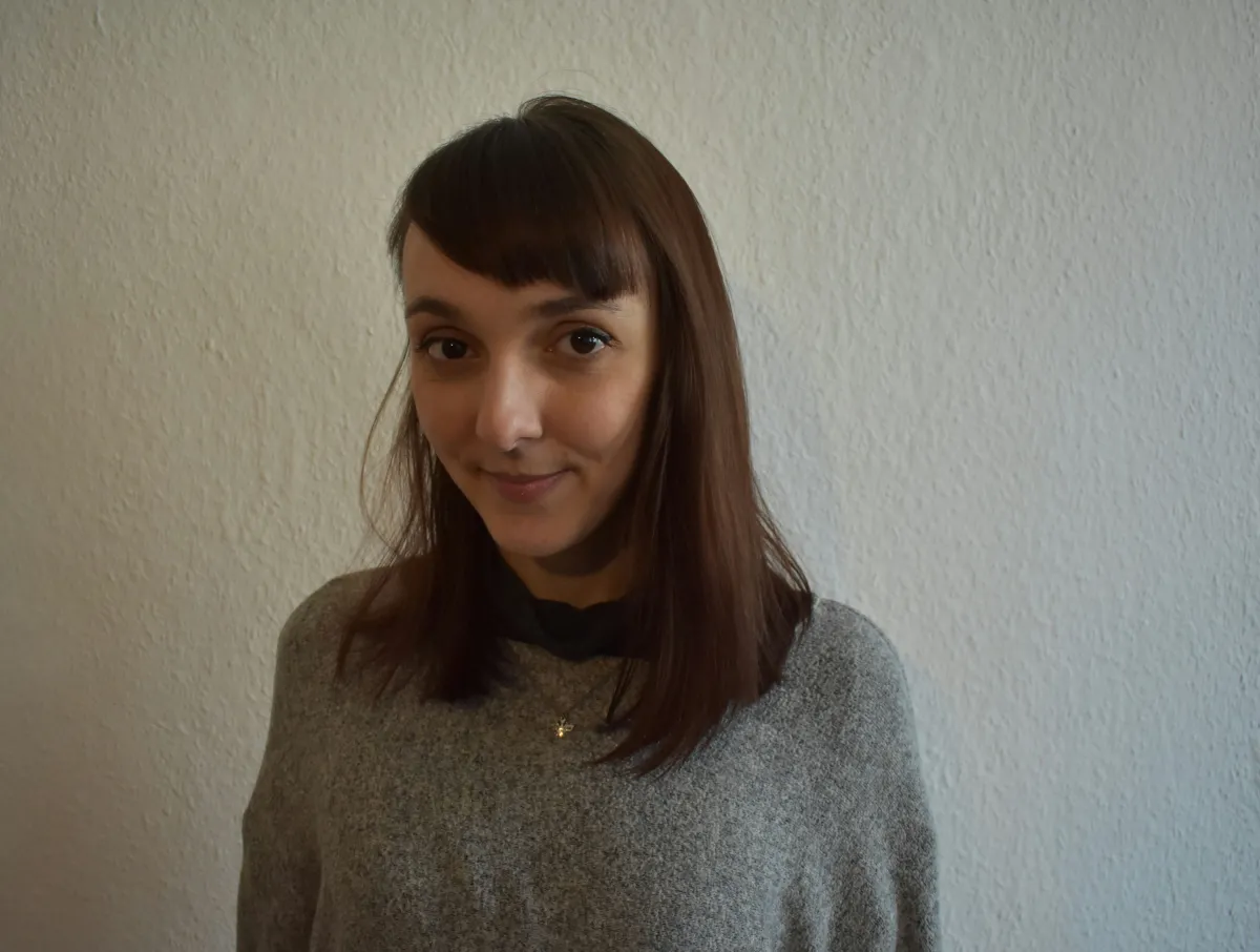 A brunette woman in a gray T-Shirt with bangs looks at the camera.