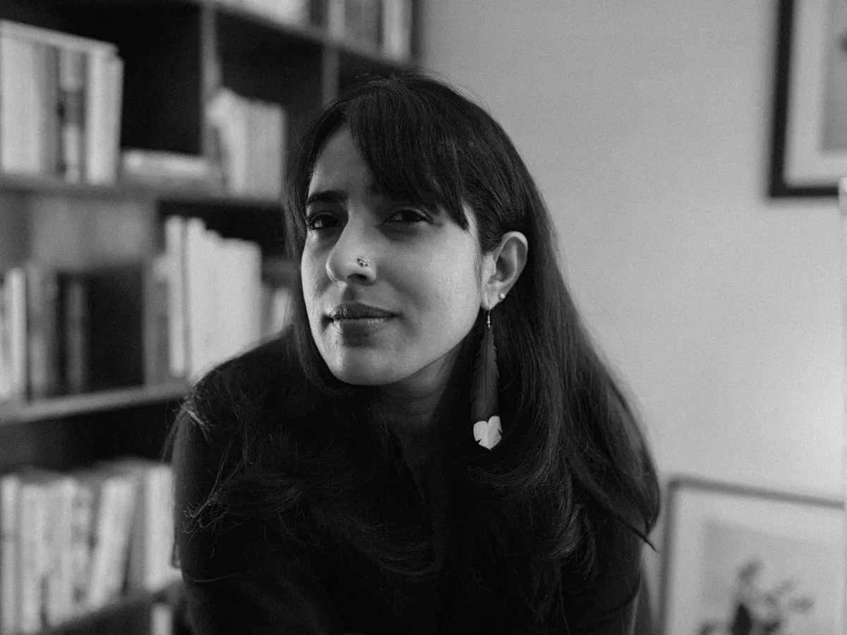 Black and white photograph of an Indian woman with mid-length hair and a long earing, sitting in front of a bookcase.