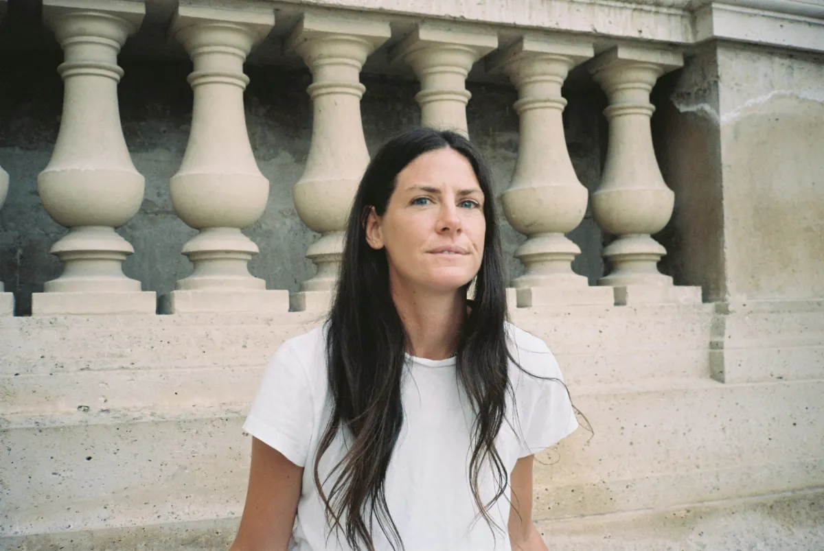 A woman with long brown hair stands in a white T-Shirt, looking at the camera.