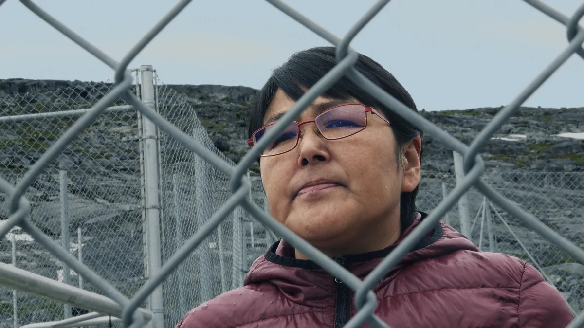 Through a chain link fence, a woman with red glasses and a red puffy jacket looks at the camera.