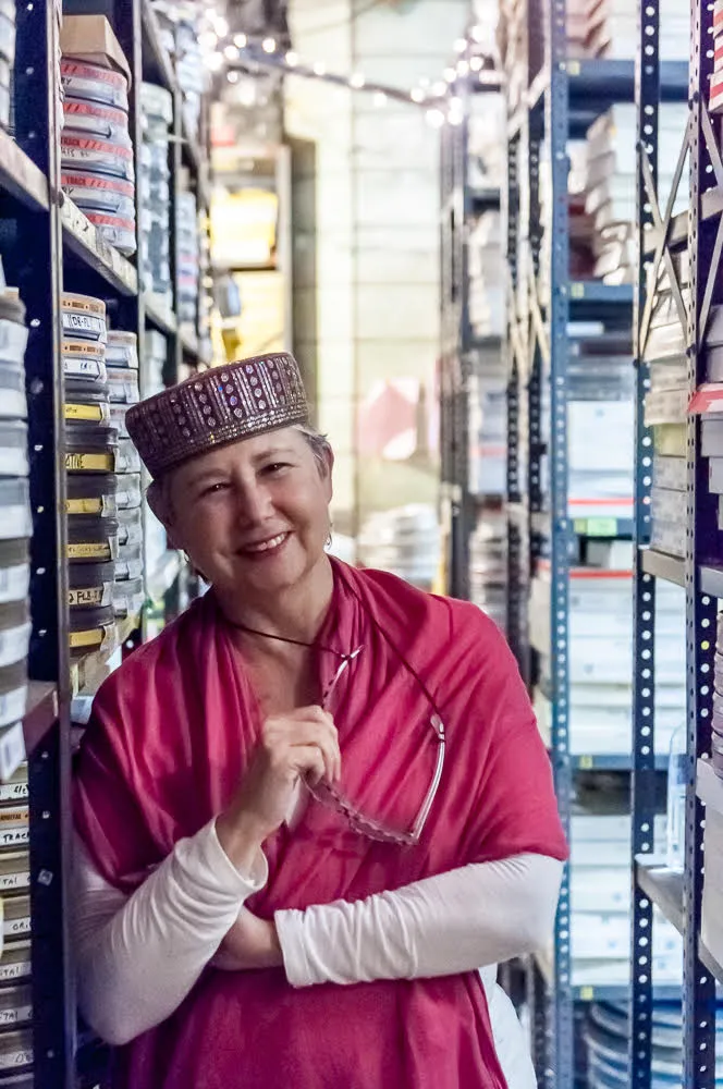 Photo of Sandra Schulberg in what looks like a film reel storage room, posing with glasses on in her right hand, wearing a pink scarf over her white longsleeve shirt and sporting a hat.