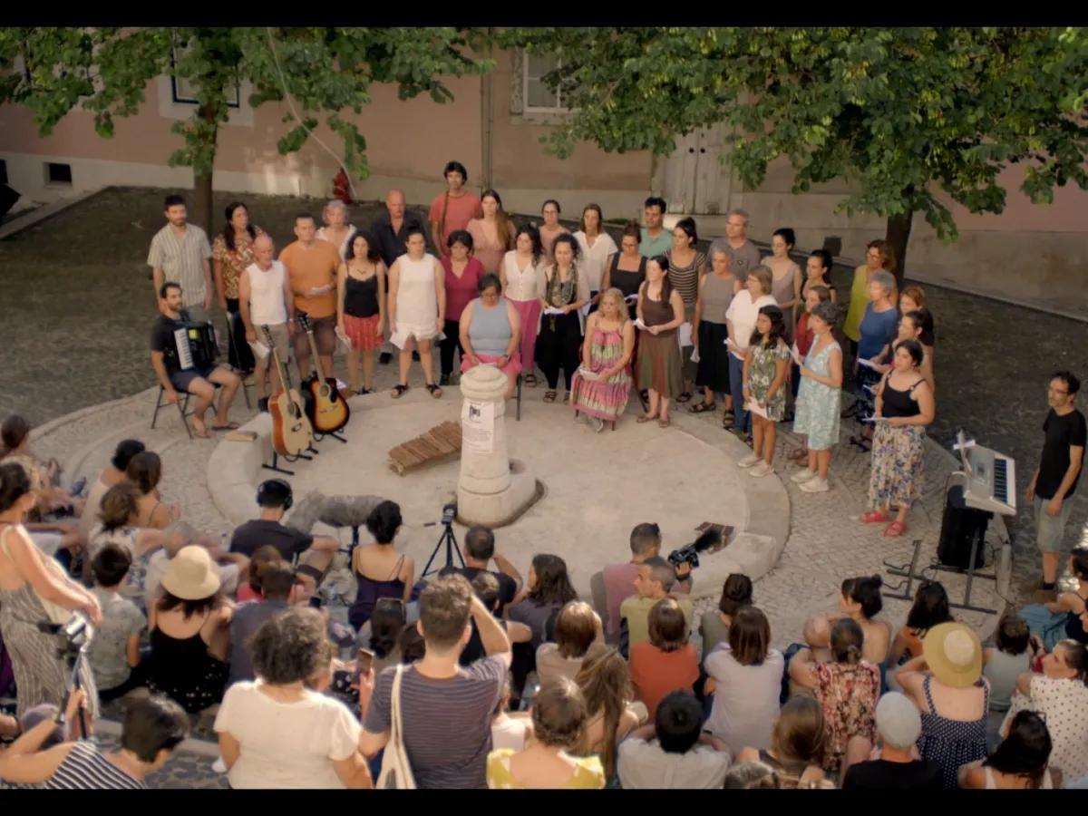 Around a public fountain, a large group of people watch a large band and choir perform.