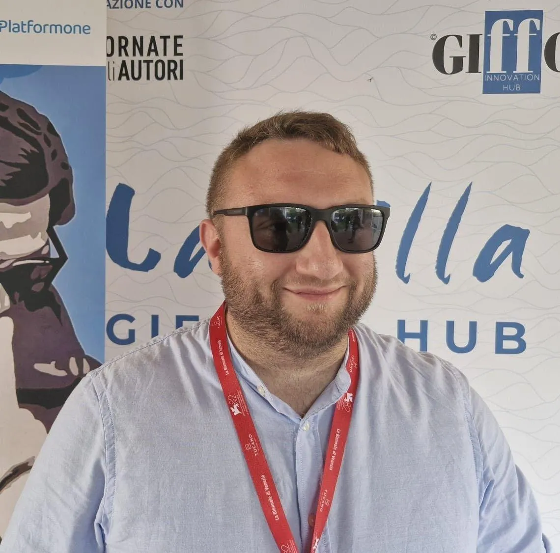 A white man wearing sunglasses smiles in front of a step and repeat.