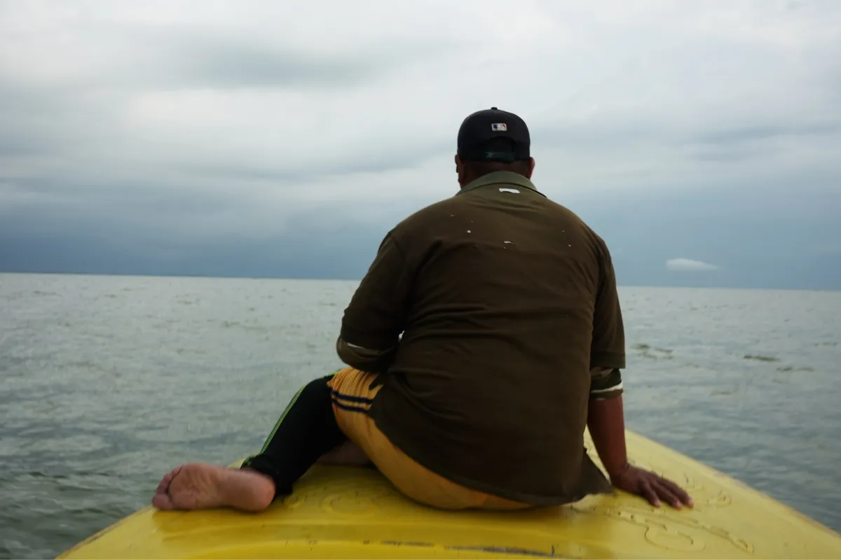 Barefoot and steady, a man sits on the bow of a yellow boat, his back to the camera, shirt weathered by time. He faces the horizon as the vessel cuts quietly through the water. The lake stretches endlessly under heavy clouds, evoking both solitude and endurance.