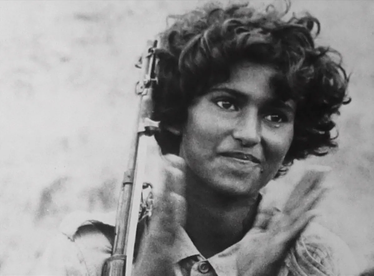 A woman smiles, while clapping, with a rifle propped against her shoulder.