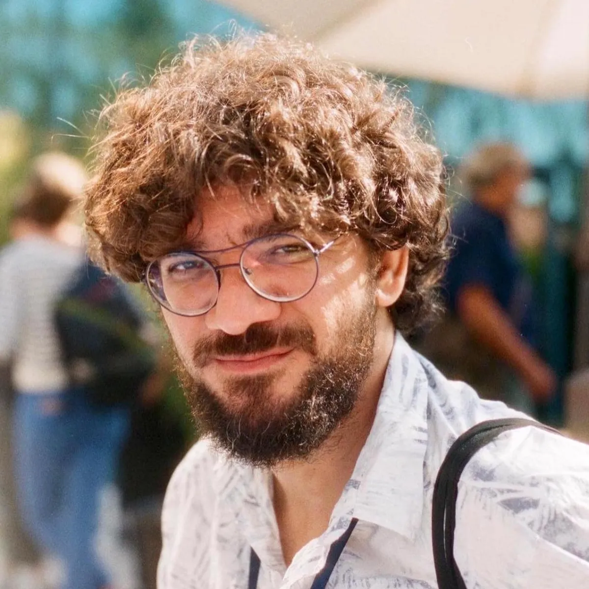 A man with curly brown hair and glasses stands in a sunny courtyard.