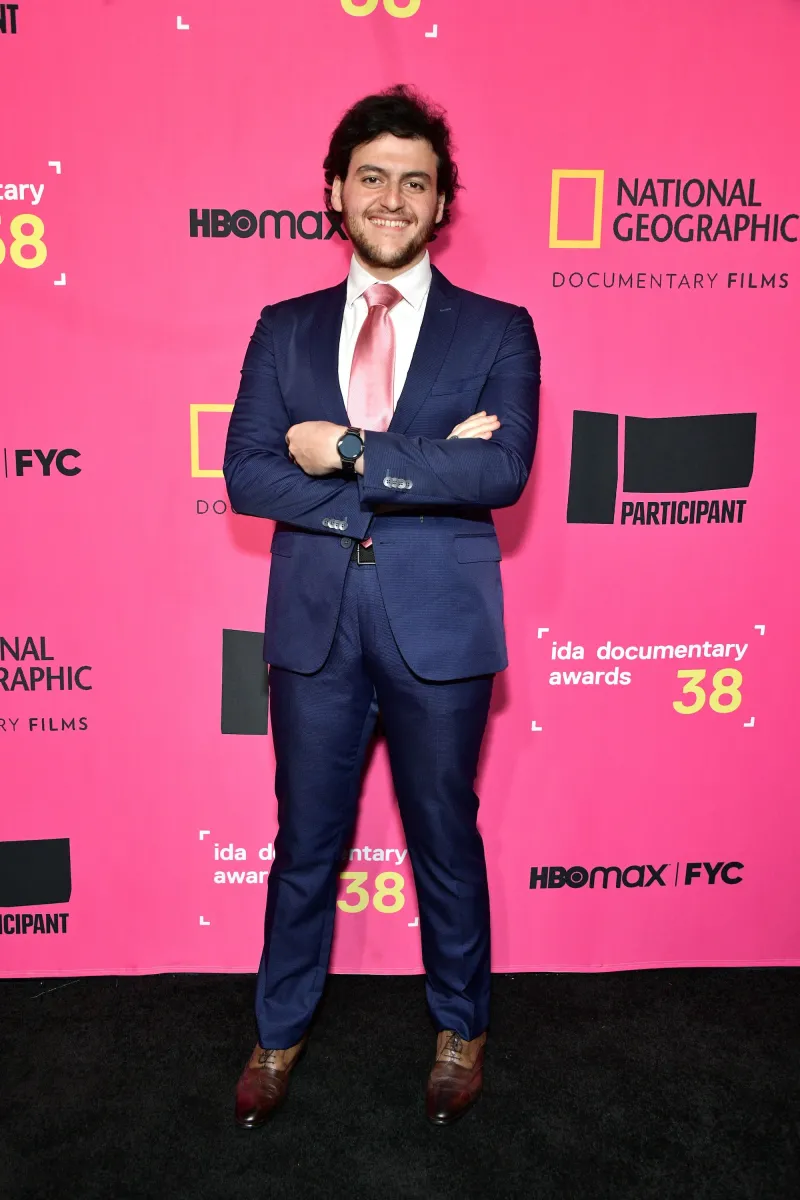 Against a pink "IDA documentary awards 38" step and repeat banner, a man in a blue suit stands with arms crossed confidentally.