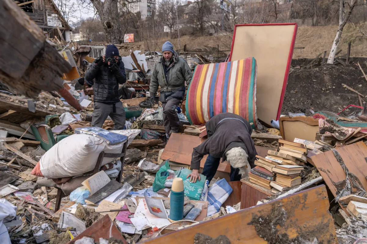 A man films another man digging through a pile of destroyed furniture and rubble.