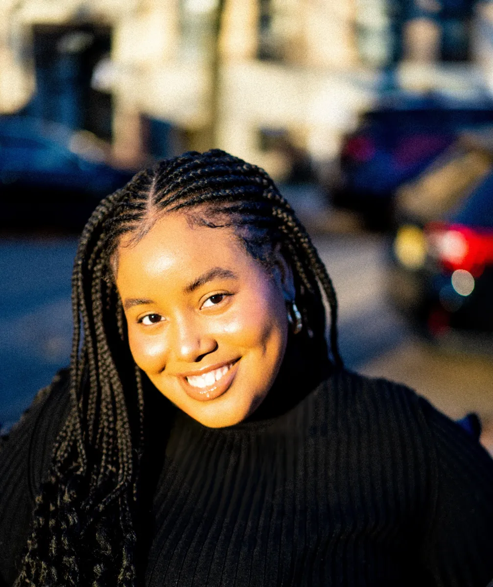 A Black woman with braided long hair smiles, with the setting sun illuminating her face.