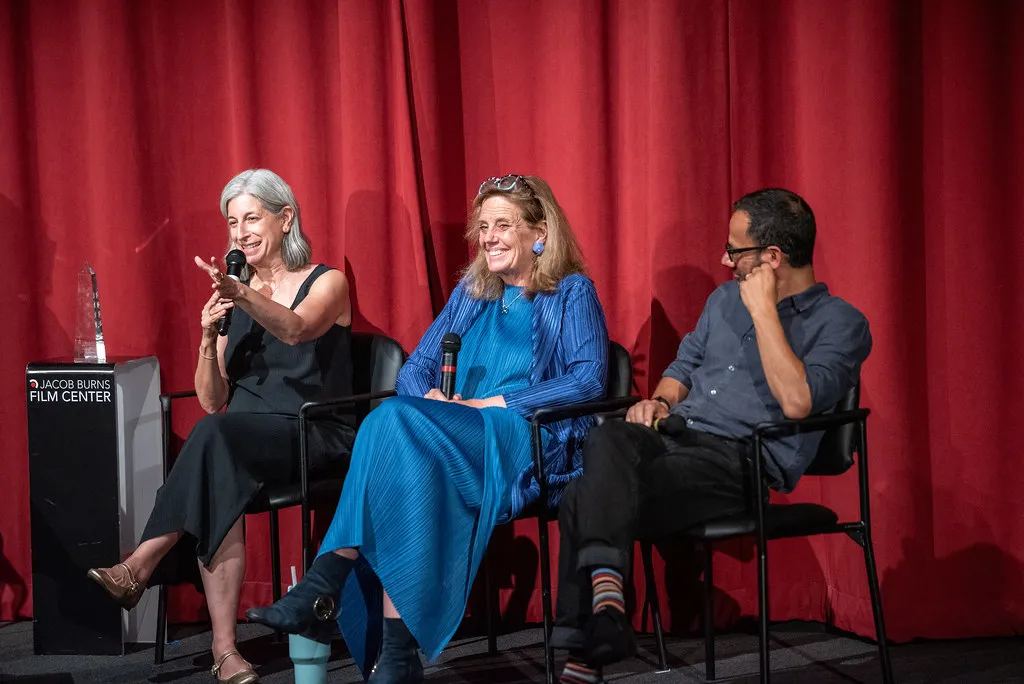 In front of red curtains, three people sit on chairs on a stage.