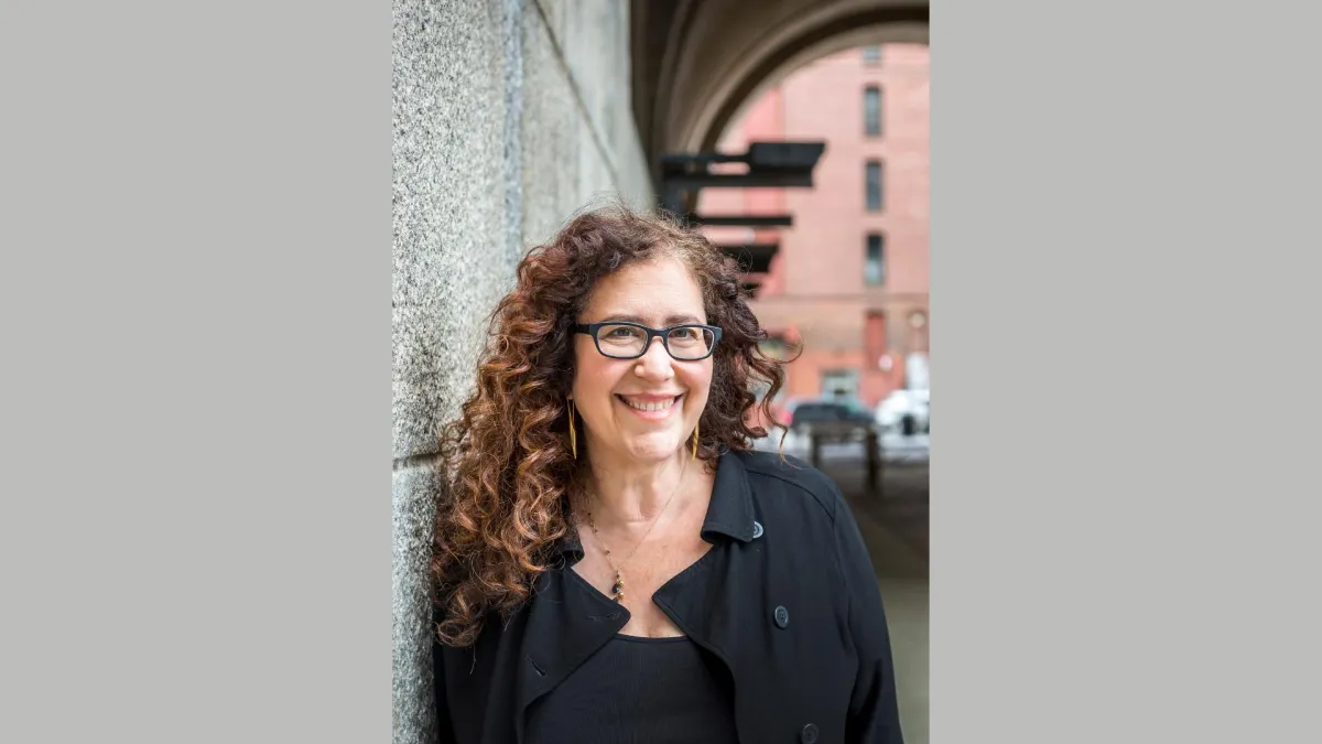A vertical image of a smiling white woman with long curly brown hair, wearing glasses and a black shirt, leaning against a stone wall.