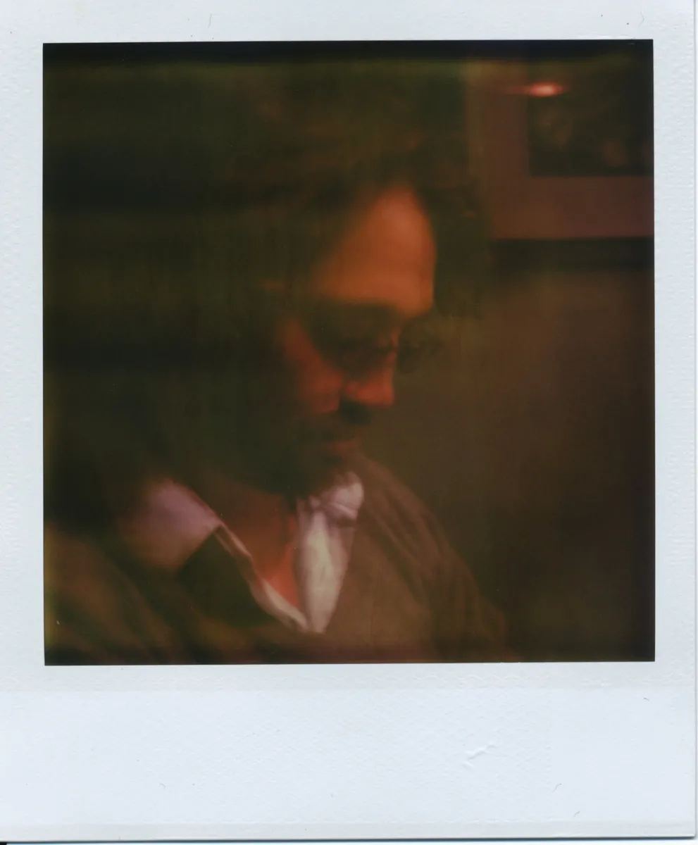 A dark, blurred polaroid of a man with curly hair and glasses wearing a white shirt and a green sweater