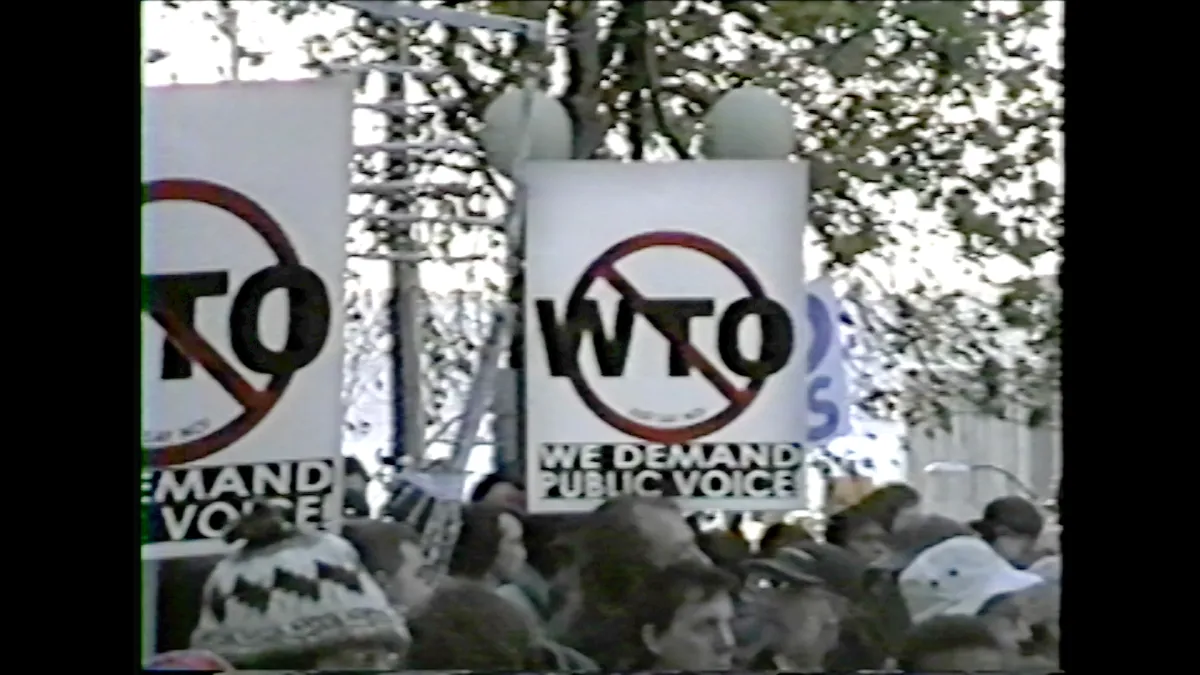 Grainy shot of group of outdoor protestors holding "We Demand Public Voice" signs with WTO crossed out
