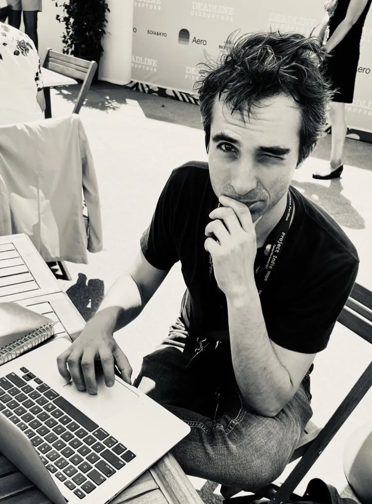 Black and white image of a light-skinned man with short dark hair wearing a tshirt and jeans sitting at a table with his laptop