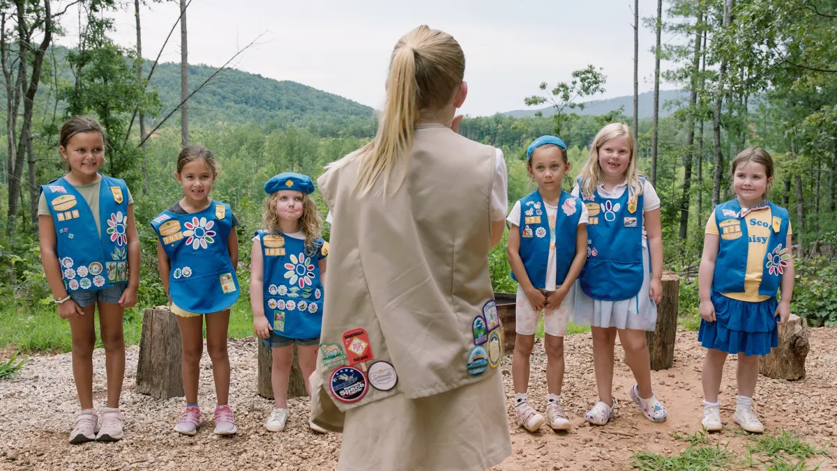 Out in a lush green forest, six young girl scouts in blue uniforms look at an older blond girl scout in a tan uniform whose back is against the camera