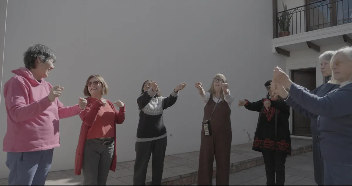 Seven women in their 70s and 80s stand in a semi-circle doing stretches in a courtyard bathed in sunlight.