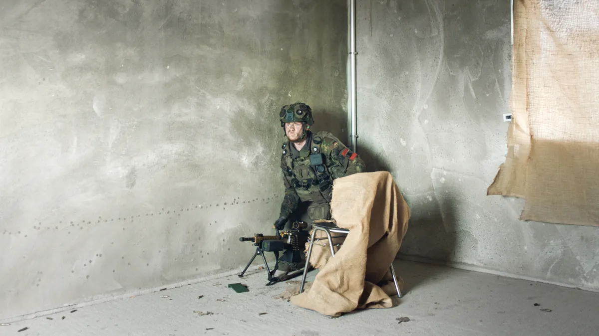 A soldier in uniform crouches inside an empty concrete room with a machine gun