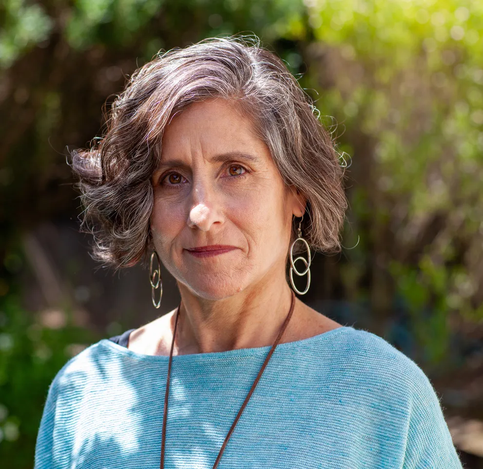 Headshot of a white woman with short brown and and white hair with long dangly earring and a baby blue shirt taken outside