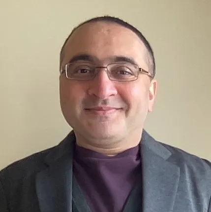 A light brown-skinned man with short black hair and glasses smiles at the camera, while wearing a grey suit and a purple t-shirt.