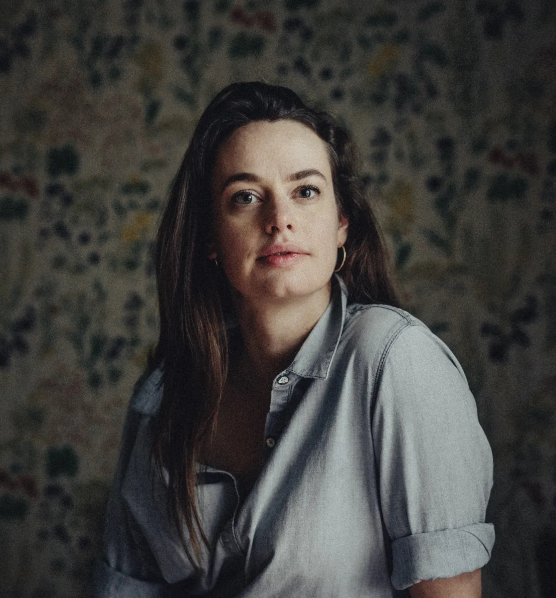 Headshot of a white woman with long dark brown hair wearing a grey button down against a floral wallpaper