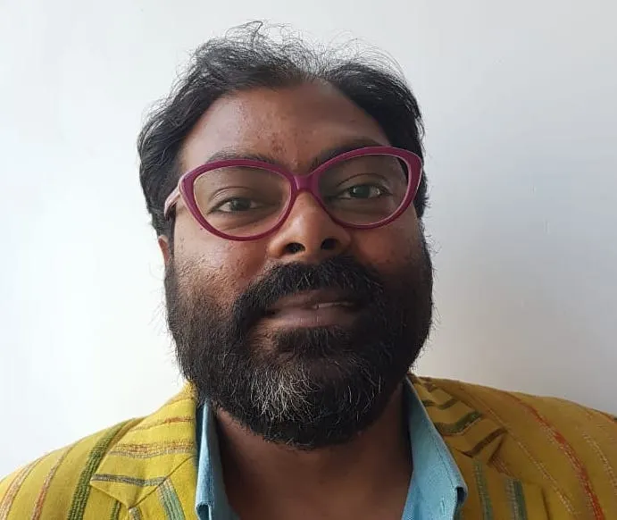 Headshot of a brown-skinned man with dark hair and bushy beard bears a bright yellow blazer, a baby blue shirt and red-rimmed glasses.