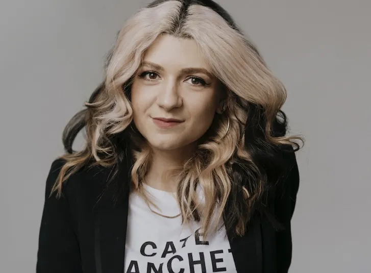 A white woman with long, dark, wavy hair dyed blond at the front wears a black jacket and a white tee that reads "Cate Blanchett"