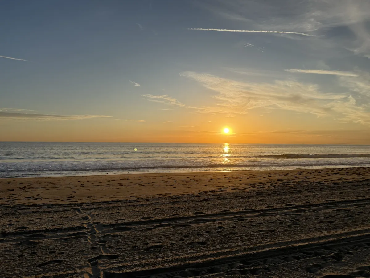 A Malibu beach at sunset with the sun over the ocean and gentle waves along the shore.