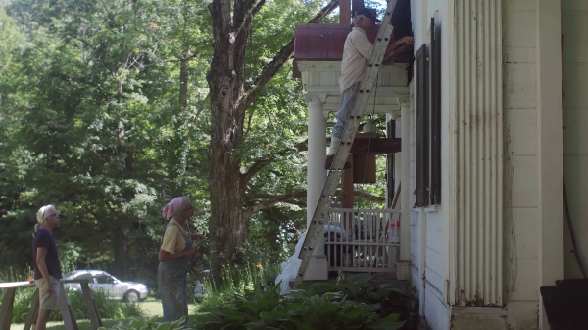 Two female sisters in their 60s and 70s look on as their brother fixes the siding of an old white house on a ladder.
