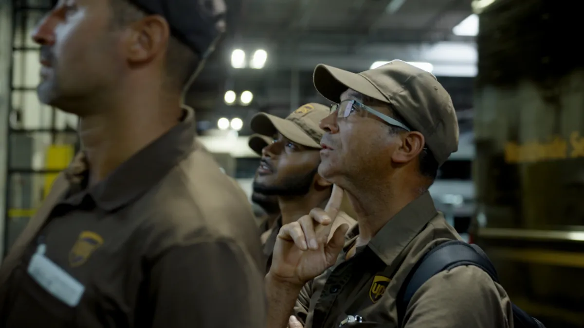 A group of men dressed in brown UPS uniforms look up, while standing inside a warehouse.